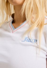 Pessoa loira a usar uma camisa branca com gola às riscas azul e rosa e texto bordado "Juicy" azul no peito.