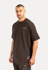 Brun t-shirt med rund halsudskæring, korte ærmer og et lille logo på brystet. Leveres med et mærke nær kanten.