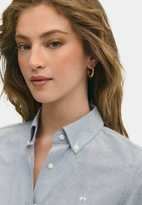 Světle modrá košile na knoflíky s límečkem, jemnou strukturou a vyšitým emblémem na levé straně; obsahuje bílé knoflíky a má přiléhavý střih.