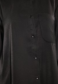 Schwarzes Hemd mit Knopfleiste aus glänzendem Stoff, das eine einzelne Brusttasche und sechs passende Knöpfe entlang der Vorderleiste aufweist.