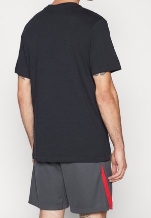 Mann trägt ein schlichtes schwarzes T-Shirt mit kurzen Ärmeln und graue Sportshorts mit rotem Streifen, von hinten gezeigt.