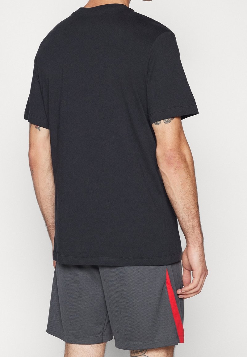 Homme portant un t-shirt noir uni à manches courtes et un short de sport gris avec une bande rouge, vu de dos.