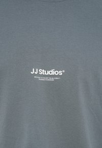 Szürke pamut póló kerek nyakkivágással. Az elején fehér nyomtatott szöveg található: „JJ Studios” és további részletek alatta.