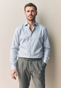 Uomo in piedi contro un muro beige, indossa una camicia azzurra a quadretti e pantaloni grigi a pieghe, con una mano nella tasca.