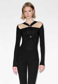 Vrouw die een zwart top met lange mouwen draagt met uitgesneden schouders en gedraaide zilveren accenten, gecombineerd met strakke zwarte broek, staand tegen een witte achtergrond.