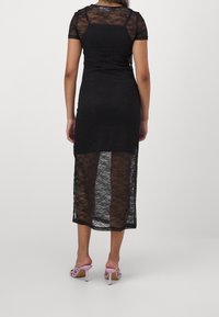 Kvinna klädd i en svart spets-middklänning med ett genomskinligt nedre kjoldel och korta ärmar, parat med rosa högklackade sandaler med öppen tå.