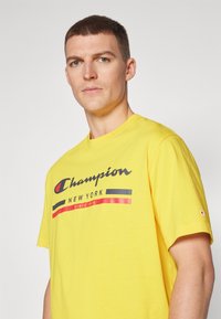 Camiseta de algodón amarilla con un logo frontal en negro y rojo que dice "Champion NEW YORK" y "SINCE 1919". Tiene mangas cortas y un corte relajado.