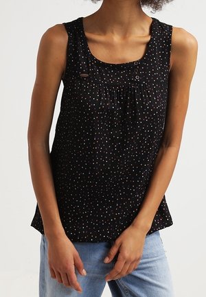 Femme portant un haut noir sans manches avec de petits points multicolores, associé à un jean bleu clair, les mains légèrement croisées à la taille.