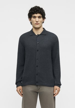 Άντρας με σγουρά μαλλιά που φοράει σκουρόγριτη ζακέτα με κουμπιά και ανοιχτό καφέ παντελόνι, στέκεται μπροστά σε απλό φωτεινό φόντο.