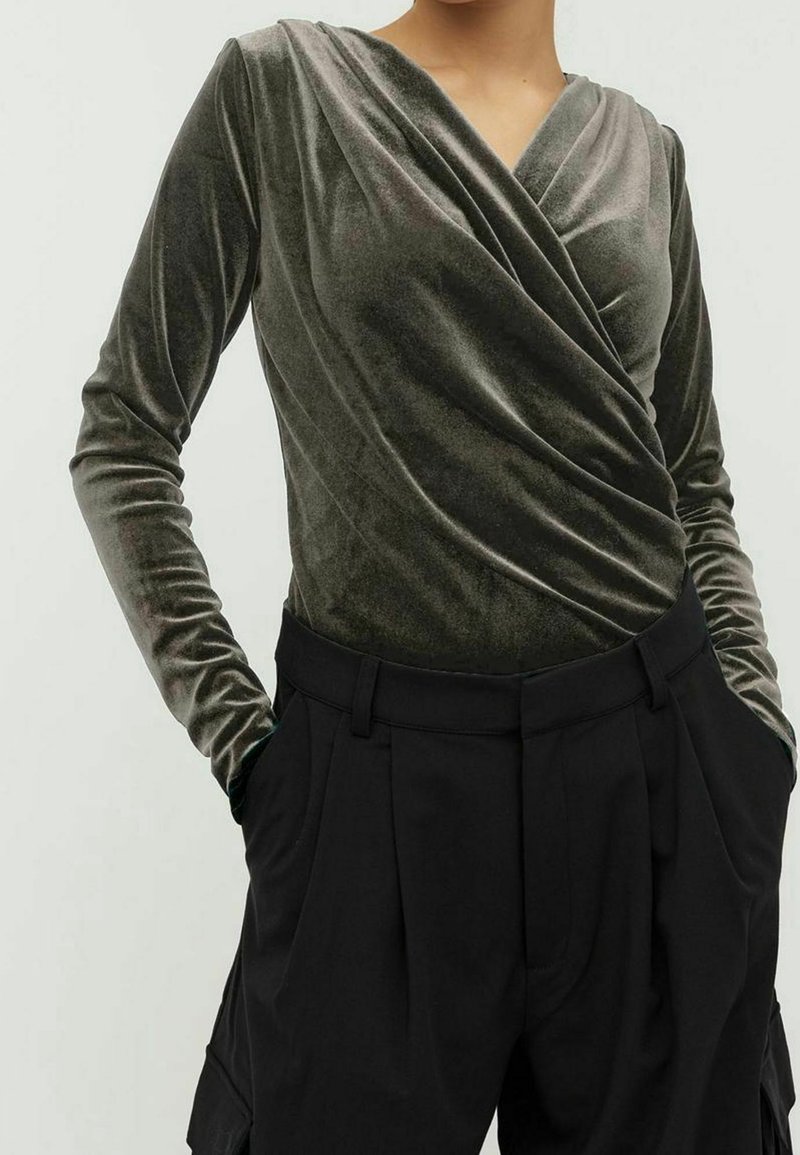 Femme portant un haut cache-cœur en velours vert olive à manches longues associé à un pantalon noir plissé taille haute, les mains dans les poches.