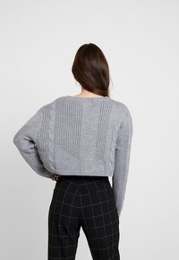 Szary krótki sweter z fakturowanym prążkowaniem i wzorami warkoczy, z szerokim okrągłym dekoltem i luźnym krojem, zestawiony z czarnymi spodniami w kratę.