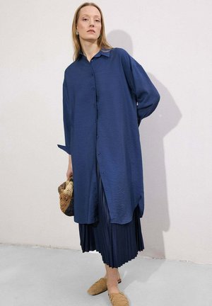 Γυναίκα που φορά μακρύ μπλε πουκάμισο με κουμπιά πάνω από μια πλισέ σκούρα μπλε φούστα, κρατώντας μια καφέ και μαύρη υφασμάτινη τσάντα χειρός, φορώντας καφετί πλεκτά επίπεδα παπούτσια.