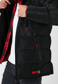 Chaqueta acolchada negra con acabado brillante, costuras horizontales, detalles en rojo, bolsillo delantero con cremallera y parche con el logo BRT en el dobladillo.