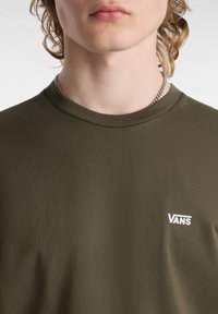 Camiseta de manga corta verde oliva hecha de tela suave, con un logo blanco de "VANS" en el área del pecho izquierdo. Cuello redondo.