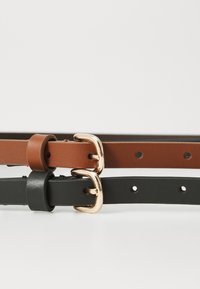Deux ceintures en cuir, l'une marron et l'autre noire, avec des boucles en métal doré sur une surface blanche.