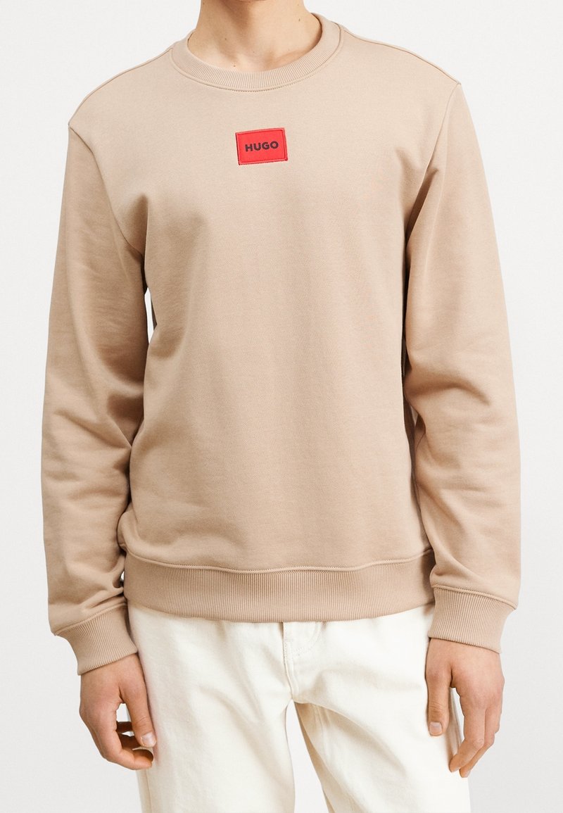 Felpa beige con scollatura a girocollo. Presenta un'etichetta logo rettangolare rossa sul petto. Realizzata in tessuto morbido con polsini e orlo a costine.