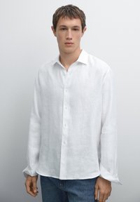 Weißes Leinenhemd mit langen Ärmeln, Knopfverschluss vorne, geradem Kragen und leicht lockerer Passform. Modell trägt blaue Jeans.