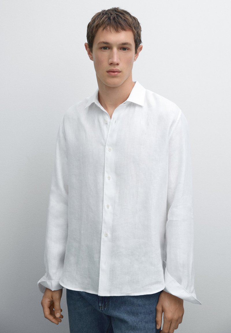 Weißes Leinenhemd mit langen Ärmeln, Knopfverschluss vorne, geradem Kragen und leicht lockerer Passform. Modell trägt blaue Jeans.