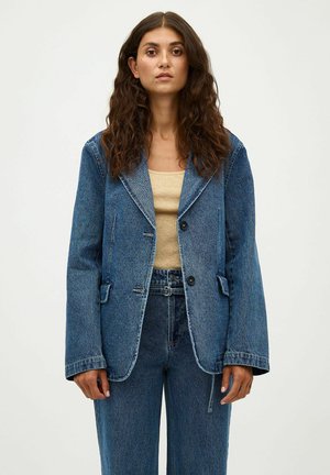 Femeie cu păr lung și creț, purtând un blazer din denim albastru și blugi de talie înaltă asortați, top bej cu dungi, stând în fața unui fundal alb.