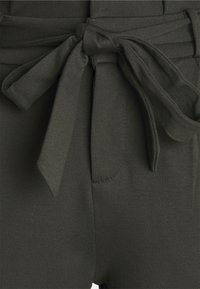 Olivgrön tygmidja med en knuten rosett, med en mjuk yta och subtil sömnad på sidorna. Inkluderar en liten ficka.