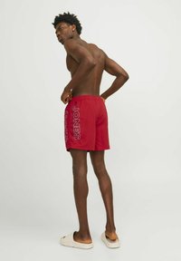 Bañador rojo con "JONES" impreso en blanco en el lado izquierdo, con cinturilla elástica y tejido ligero; acompañado de sandalias ligeras.