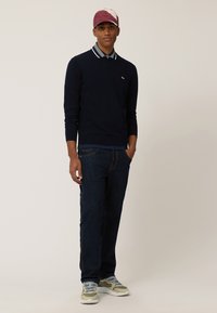 Marineblaues Sweatshirt mit langen Ärmeln und Polokragen über einem hellblau karierten Hemd, kombiniert mit dunkelblauen Jeans und beigen Sneakers.
