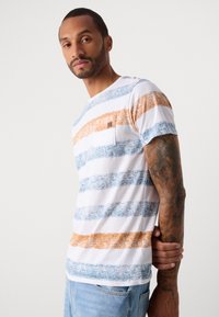 Mann med arm tatovering iført hvit t-skjorte med blå og oransje horisontale striper og lyseblå jeans, stående mot en ensfarget bakgrunn.