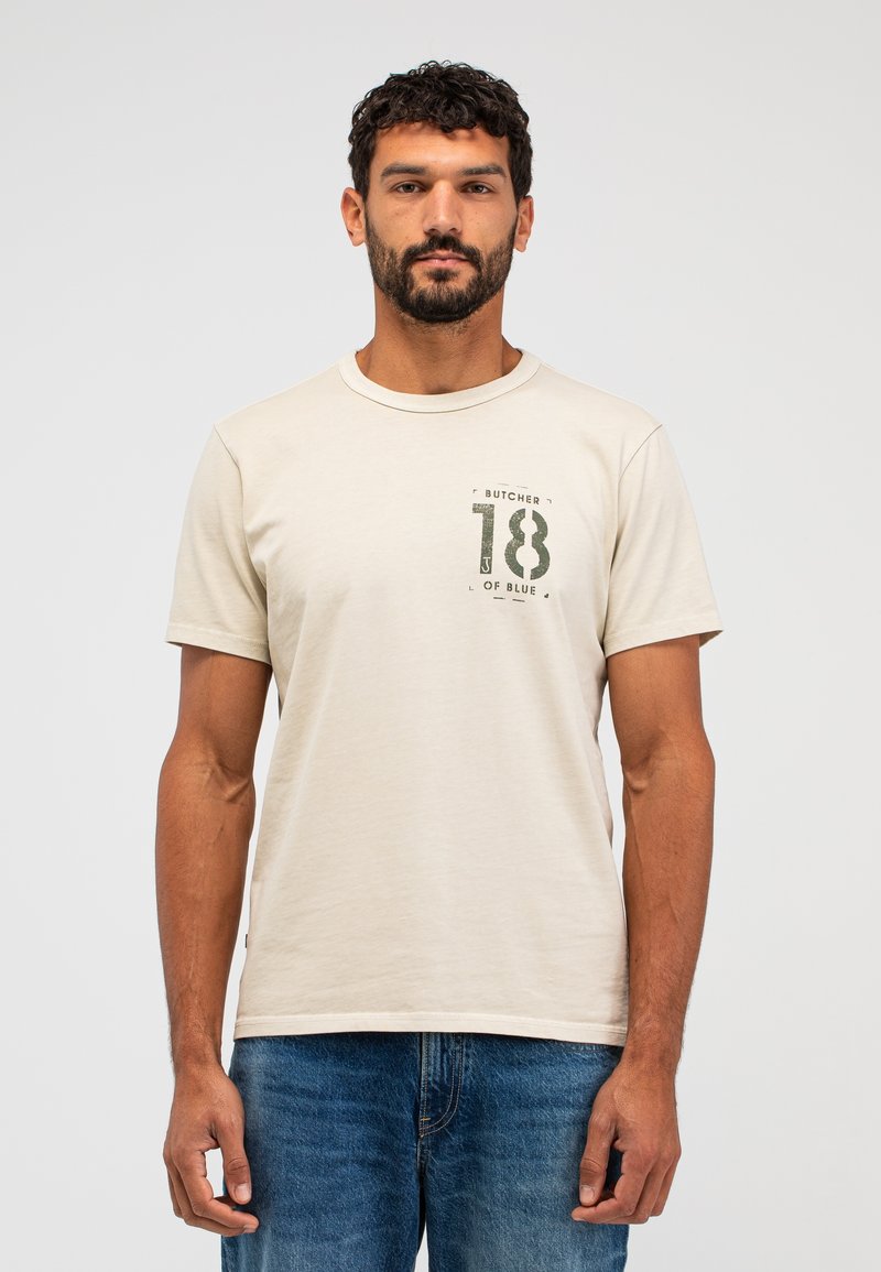 Beige T-Shirt mit kurzen Ärmeln aus weichem Baumwollstoff, mit grüner aufgedruckter Schrift "BUTCHER 18 OF BLUE" im Bereich der linken Brust.