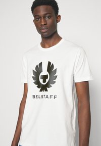 Balta kokvilnas t-kreklu ar melnu logo un spārniem, uz kura ir rakstīts "BELSTAFF". Klasisks piegriezums ar īsām piedurknēm un apaļu kakla izgriezumu.