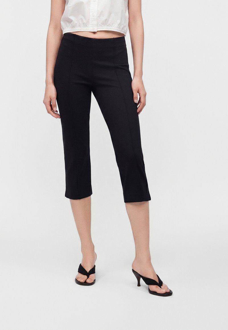 Femme porte un pantalon noir raccourci et des sandales noires à talons avec un crop top blanc sans manches à boutons, debout sur un fond uni.