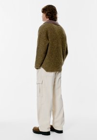 Oliivinvihreä neulepaita vaaleanpurppuraisella kauluksella, yhdistetty kermaisiin cargo-housuihin, joissa on sivutaskut ja rento leikkaus. Mustat kengät.