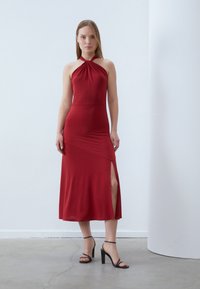 Red halter-neck dress with a fitted bodice, asymmetrical skirt, and side slit. Made of a smooth fabric, features a minimalist design.