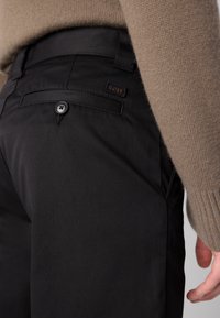 Sorte bukser lavet af glat stof, med en baglomme med knaplukning og et lille brunt logo mærke. Blød brun sweater synlig.