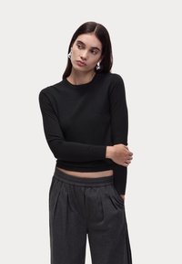 Top lungo a maniche lunghe nero in tessuto morbido, abbinato a pantaloni grigi plissettati con cintura elastica e tasche laterali.