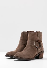 Botas de ante marrón a la altura del tobillo con punta redonda, hebillas laterales y un tacón de bloque. Presentan una suela texturizada y costuras decorativas.