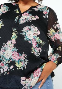 Femme portant un chemisier noir à manches longues avec des motifs floraux colorés et un tissu texturé, main dans la poche, associée à un jean bleu.