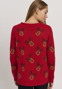 Pull en laine rouge avec un motif de têtes de renne, ornées de cornes brunes et de visages beiges, avec un col et des poignets côtelés.
