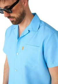 Camicia a maniche corte azzurro chiaro realizzata in tessuto morbido, con taschino, chiusura a bottoni e un'etichetta arancione come accento sulla tasca.