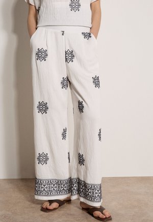 Weiße, weit geschnittene Hose mit schwarzer Blumenstickerei, getragen mit den Händen in den Taschen und kombiniert mit braunen, offenen Sandalen auf einem beigen Boden.