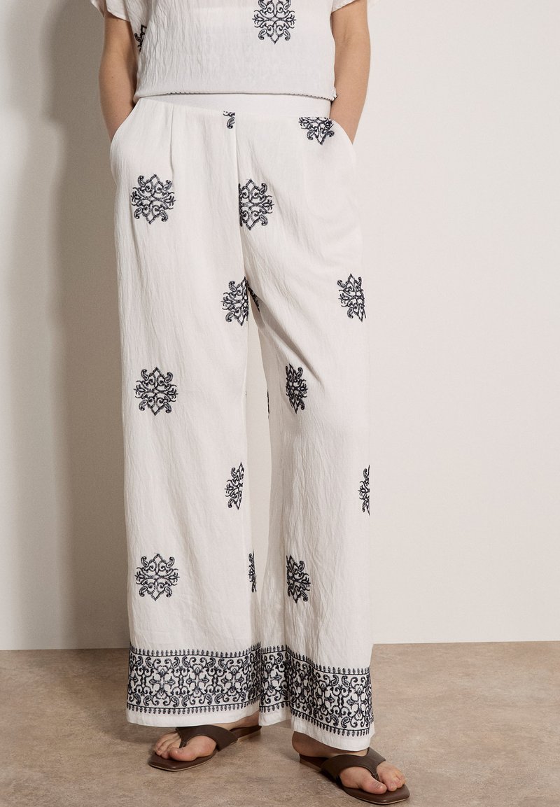 Pantalon blanc à jambes larges avec broderie florale noire, porté les mains dans les poches et associé à des sandales marron à bout ouvert sur un sol beige.