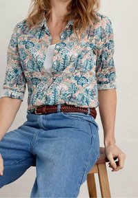 Geknöpftes Hemd mit Muster und weißem Innenfutter, versehen mit blauen und grünen Pflanzenmotiven auf einem pfirsichfarbenen Hintergrund, kombiniert mit hochtaillierten Jeans.