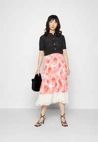 Cardigan preto de malha com mangas curtas e detalhes de botões, combinado com uma saia plissada em rosa e branco com padrão floral. A modelo segura uma clutch de pele preta.