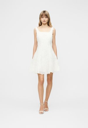 Mladá žena stojící, oblečená v bílé květinové texturované šaty bez rukávů a bílé boty na podpatku s páskem u kotníku, na jednobarevném světlém pozadí.