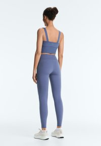 Femme debout, de dos, portant un soutien-gorge de sport bleu et des leggings taille haute avec des baskets blanches sur fond blanc.