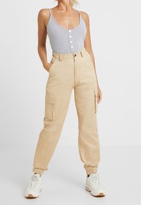 Grijze bodysuit met knopen, beige cargo-broek met zakken, taps toeloopende boorden, en witte sneakers met bruine accenten. Een schone, casual uitstraling.