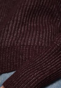 Matière de pull côtelé bordeaux avec un tricot texturé, présentant un gros plan de la manche sur un denim bleu clair.
