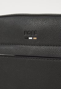 Black textured leather bag with embossed "BOSS" logo. Features three colored dots: black, white, and brown. Clean, minimalist design.