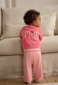 Pull en tricot rose avec "SMILE" brodé en crème et un cœur, associé à un pantalon rose côtelé. L'ensemble présente des accents volants.