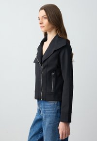 Zwarte, aansluitende jas met een geknipte kraag, ritssluiting aan de voorkant en twee ritssluitingenzakken. Gemaakt van soepel, flexibel materiaal met een nette afwerking.
