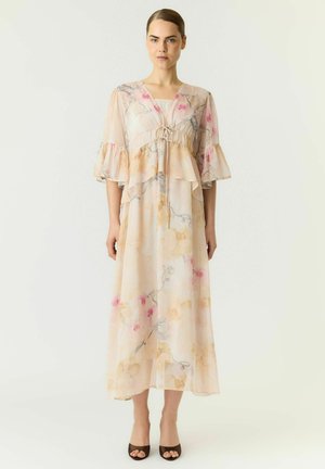 Femme portant une longue robe fleurie rose clair avec des manches à volants et un lien au niveau de la poitrine, debout sur un fond blanc uni.